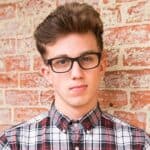 Young person wearing black glasses and a plaid shirt stands in front of a brick wall, looking at the camera.