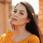 A person with long dark hair and wearing an orange top is looking into the camera, with one hand touching their face. An outdoor scene with blurred background.